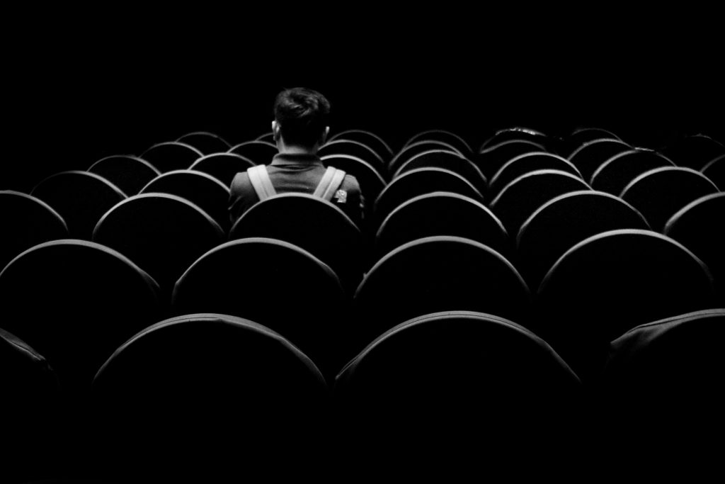Ein Mann sitzt allein im Publikum in einem dunklen Theater, umgeben von leeren, schwarzen Stühlen, die sich in wiederholenden Mustern erstrecken.